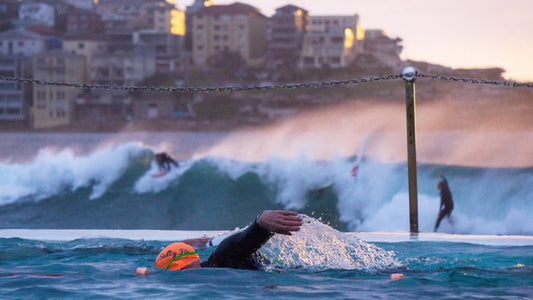 swimmer in outdoor pool with surfers in the sea behind 
