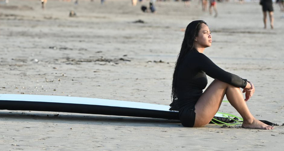 a lady wearing a shorty wetsuit sat on a sandy beach with a longboard next to her